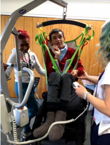Salih is excited about trying out a standing frame. He is listening carefully to the OT and PT who are helping him know how to move and sit in the hoist on the way to try the frame.